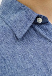 Camisa de lino azul con cuello abotonado, tejido texturizado y un solo botón en la parte delantera. Ligera y con un diseño casual.