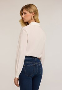 Blusa a maniche lunghe di un beige chiaro con colletto e polsini con bottoni, abbinata a jeans a vita alta di un blu scuro. Tessuto dal tocco morbido.