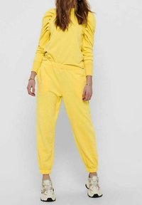 Conjunto de sudadera amarilla y pantalón jogger a juego, tela suave, puños y cintura elásticos, mangas ligeramente abullonadas, combinado con zapatillas beige y negras.