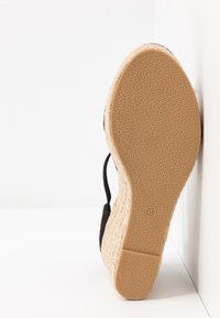 Schwarzer Schuh mit strukturiertem Obermaterial, Keilabsatz aus Jute und detaillierter Gummisohle. Der Schuh ist vor einem weißen Hintergrund platziert.