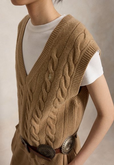 Person iført en beige kabelstrikket ærmeløs sweater over en hvid skjorte, kombineret med tanfarvede bukser og et bredt brunt bælte med udsmykkede sølvspænder.