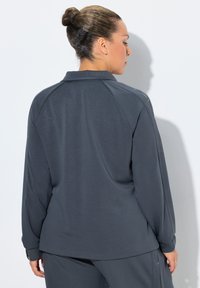 Mujer con el cabello recogido en un moño, vistiendo una camisa de manga larga gris oscuro y pantalones a juego, de pie frente a una pared blanca con sombra visible.