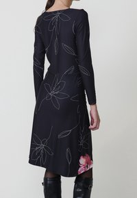 Femme portant une robe noire à manches longues avec des motifs floraux blancs en lignes et une fleur rose près de l'ourlet, associée à des bottes noires montantes.
