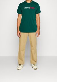 Camiseta de algodón verde con un logo blanco y rojo de "Tommy Jeans", combinada con pantalones beiges y zapatillas blancas con acentos beige.
