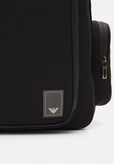 Mochila negra hecha de material duradero con una superficie texturizada, cierre de cremallera y un parche de logo rectangular que presenta un emblema estilizado.