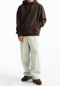 Felpa con cappuccio marrone e tasca frontale, abbinata a jeans baggy blu chiaro e sneakers marroni con superficie texturizzata.