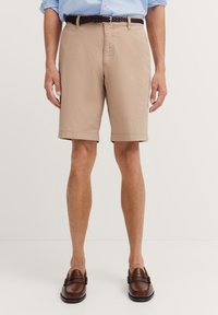 Beige Shorts aus leichtem Stoff, mit flachem Bund, Seitentaschen und einem Saum, der oberhalb des Knies endet. Brauner geflochtener Gürtel und Schuhe.