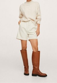 Pull en maille torsadée crème, short beige et bottes en cuir marron montantes jusqu'au genou avec semelle plate. Design simple, coupe structurée et texture lisse.