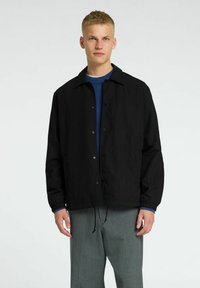 Giacca leggera nera con bottoni a pressione, tasche laterali e polsini elastici, indossata sopra un maglione in maglia blu. Indossata con pantaloni grigi.