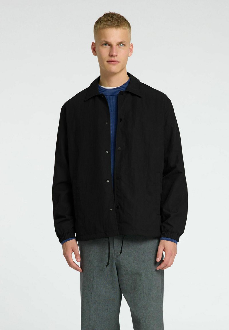 Giacca leggera nera con bottoni a pressione, tasche laterali e polsini elastici, indossata sopra un maglione in maglia blu. Indossata con pantaloni grigi.