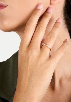 Frau berührt mit der linken Hand die Wange, trägt einen goldenen Ring mit kleinen weißen Perlen am Mittelfinger, hellrosa Nagellack, dunkle grüne Kleidung sichtbar.
