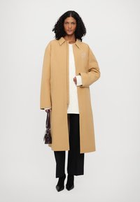 Lys brun frakke med krave-design, skrå front og sidelommer, parret med en hvid struktureret sweater og sorte vide bukser.