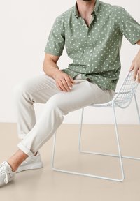 Koszulka zielona z krótkim rękawem w biały diamentowy wzór, w połączeniu z jasnobeżowymi spodniami i białymi trampkami, siedząca na jasnoniebieskim metalowym krześle.