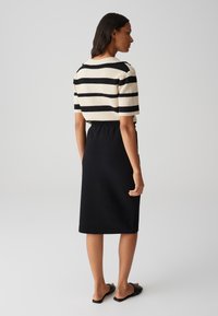 Strickoberteil mit schwarzen und cremefarbenen Streifen, kombiniert mit einem schwarzen Midil裙, der über einen elastischen Bund verfügt. Flache Sandalen vervollständigen den Look.