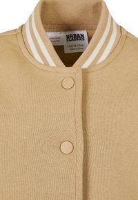 Beige bomberjakke med ribbet krave med hvide striber, frontknapper og en glat tekstur. Mærkeetiketten er synlig indeni.