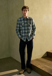 Camisa de cuadros en azul y gris, con dos bolsillos en el pecho; combinada con jeans oscuros y zapatos marrón claro. De pie en unas escaleras de madera con una pared texturizada.