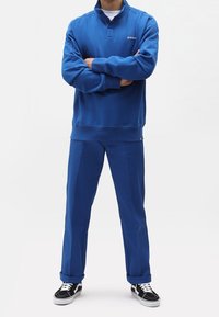 Modrý fleecový pulovr s vysokým límcem a zapínáním na knoflíky, doplněný odpovídajícími modrými kalhotami. Černobílé tenisky s vzorem dokončují vzhled.