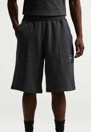 Mężczyzna noszący czarne szorty do kolan z przednimi kieszeniami i małym logo Nike na lewej nodze, stojący na jednolitym tle.