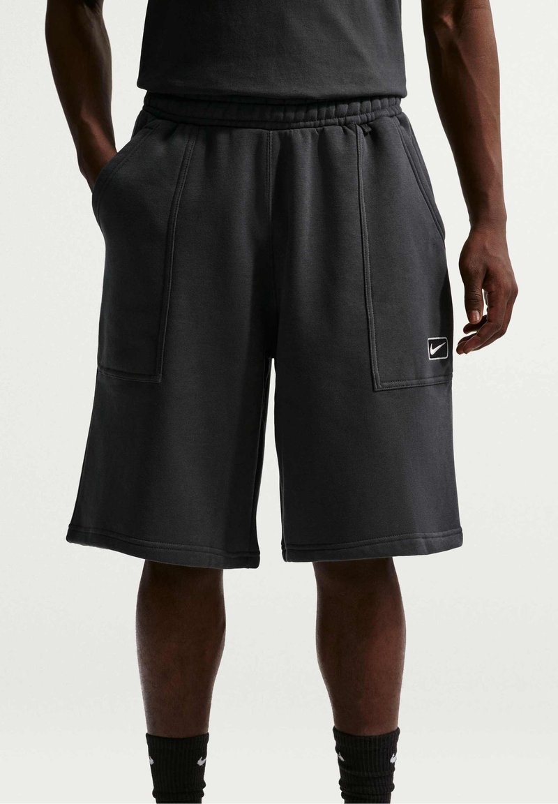 Homme portant un short noir au genou avec des poches devant et un petit logo Nike sur la jambe gauche, debout devant un fond uni.