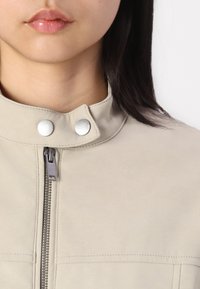 Bež jakna od umjetne kože s visokim ovratnikom, srebrnim kopčama na dugmadi i prednjim zatvaračem. Glatka tekstura s detaljem ušivanja.