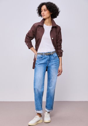 Kiharatukkainen nainen, joka pukeutunut ruskeaan takkiin, valkoiseen t-paitaan, vaaleansinisiin käärityihin farkkuihin, harmaaseen punottuun vyöhön ja valkoisiin lenkkareihin, seisoo käsi lanteilla.