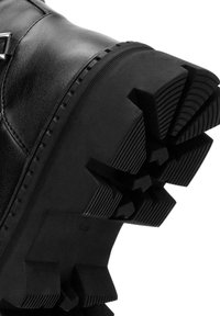 Bota de cuero negra con suela de goma texturizada que presenta un patrón de relieve pronunciado y notables estrías, adecuada para terrenos ásperos.