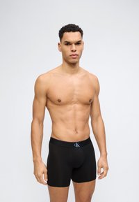 Skjorteløs mann med kort krøllete hår som står mot en ensfarget bakgrunn, iført svarte Calvin Klein-bokser.