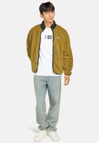 Fleece jack in olijfgroen met zwarte kraag, ritssluiting aan de voorkant en logo-patch op de borst. Gecombineerd met lichtblauwe jeans en witte sneakers.