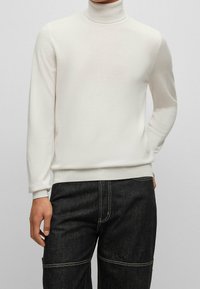 Weißer Rollkragenpullover aus weichem Material, mit langen Ärmeln und gerippten Säumen, gezeigt zu dunklen Jeans.