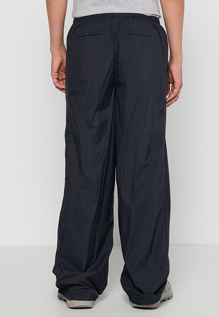 Persoon in losse zwarte nylon broek en grijze sneakers, staand tegen een effen witte achtergrond, bekeken van achteren.