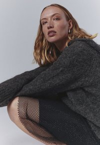 Suéter gris oscuro y texturizado con un corte holgado, combinado con una falda de red negra. Piel suave, pendientes de aro simples y cabello ondulado de longitud media.