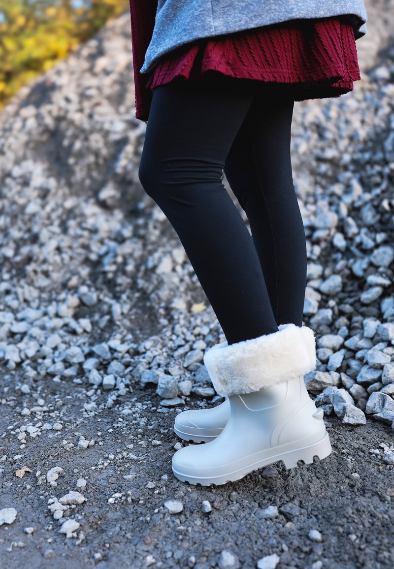 Bottes blanches imperméables avec un bord en fourrure blanche et duveteuse, portées avec des leggings noirs, debout sur un terrain de gravier.