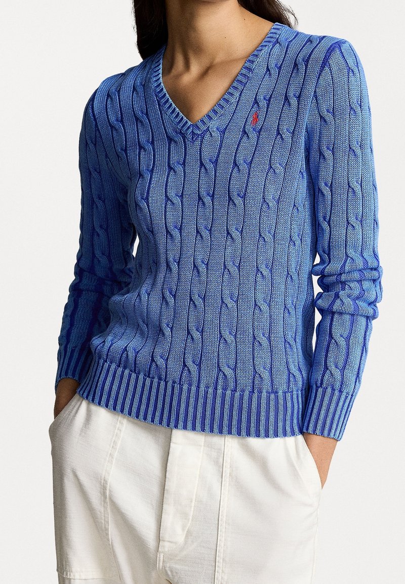 Femme portant un pull bleu à col en V avec motif torsadé, petit logo rouge sur la poitrine et pantalon blanc, mains dans les poches, fond neutre.