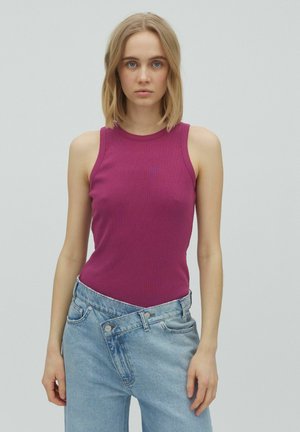 Tânără cu păr blond, purtând un top fuchsia fără mâneci și blugi bleu cu talie înaltă, cu talia nasturată descheiată.