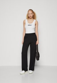 White tank top with "GAP" logo, black wide-leg pants, and white sneakers. Accessories include a small black shoulder bag. Smooth textures.
