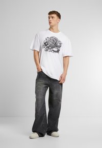 White graphic t-shirt featuring black design at the center, paired with dark gray wide-leg jeans and white sneakers on a neutral background.