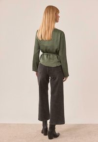 Top verde de manga larga con cintura anudada, combinado con vaqueros de pierna ancha de talle alto en gris oscuro y botines negros de tacón. Textura de tela suave.