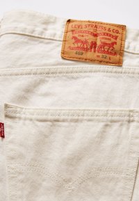 Valkoiset farkkibbleet, joissa on ruskea nahkapaikku, johon on kirjailtu "Levi Strauss & Co." punaisella ompelulangalla ja erottuvalla takataskusuunnittelulla.