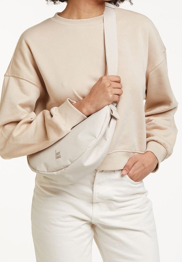 Ljusbeige axelväska i tyg med en svängd form och en diskret logotyp. Bärs över en matchande beige sweatshirt och vita byxor.