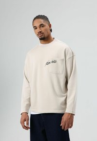 Beige sweatshirtt met lange mouwen, ronde hals en een voorzak. Voorzien van een zwart grafisch ontwerp op de zak. Zachte stoftextuur.