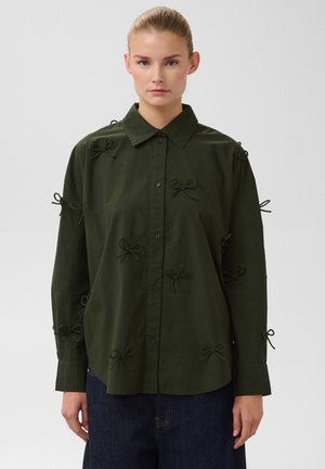 Femme aux cheveux blonds en chignon porte une chemise boutonnée vert foncé ornée de petits nœuds et un pantalon bleu foncé, debout devant un fond uni.