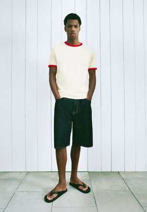 Mand står med hænderne i lommerne iført en cremefarvet T-shirt med rød kant, mørke knælange shorts og sorte klipklapper mod en lys væg.