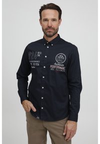 Marineblå skjorte med knappelukking og hvite logoer og detaljer, med klassisk krage og lange ermer, laget av et glatt og lett stoff.