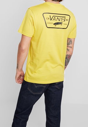 Osoba nosząca żółtą koszulkę Vans z czarnym logo i grafiką deskorolki na plecach, w połączeniu z ciemnoniebieskimi jeansami i widocznymi tatuażami na ramionach.