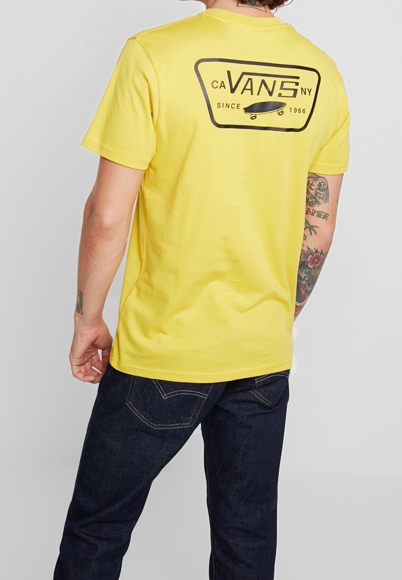 Personne portant un t-shirt Vans jaune avec un logo noir et un graphique de skateboard au dos, associé à un jean bleu foncé et des tatouages visibles sur le bras.