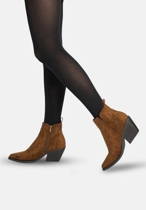 Gambe con collant neri che indossano stivaletti in suede marrone con cerniera laterale, motivi floreali traforati e tacco a blocco impilato.
