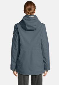 Vrouw met opgestoken haar in een blauwe hoodie, gezien van achteren tegen een effen achtergrond.