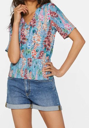 Kvinde iført en blå blomstret bluse med knapper, rynket talje og omsnoede denimshorts, den ene hånd på hoften og den anden nær hagen.