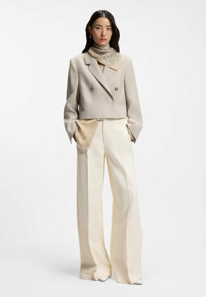 Woman wearing a beige cropped double-breasted blazer, silk patterned scarf, wide-leg cream trousers, and beige blouse with hands in pockets.