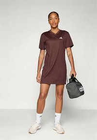 Karmiinpunane spordikleit lühikeste varrukatega, v-kaeluse disain ja valged küljeribad. Mudel hoiab musta adidas kotti. Sile kangastekstuuri.